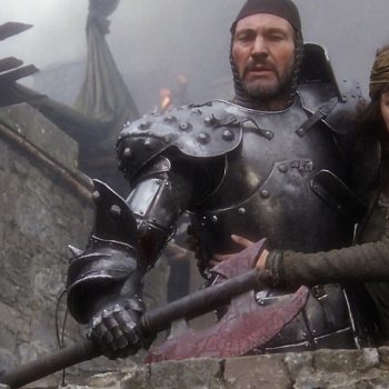 Patrick Stewart as Leodegrance and Cherie Lunghi as Guinevere in John Boorman's "Excalibur" during the siege of her father's castle. Armored knight and woman in medieval dress standing on stone castle wall in misty battle scene from 1981 film.