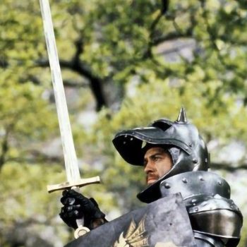 Nigel Terry as King Arthur in John Boorman's "Excalibur" embodies the legendary once and future king in full armor. Armored knight holding sword upward with dragon heraldry on breastplate in outdoor forest setting from 1981 film.